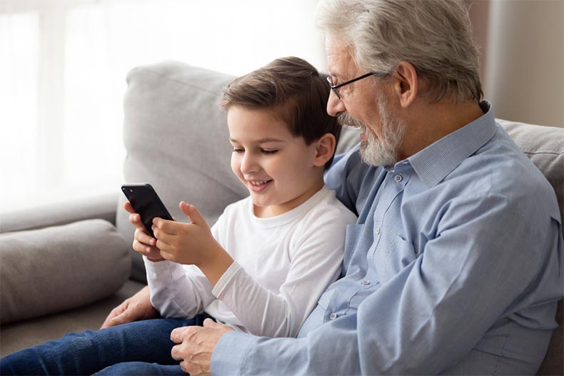 A grandfather sitting on a couch with his grandson who's holding a smart phone