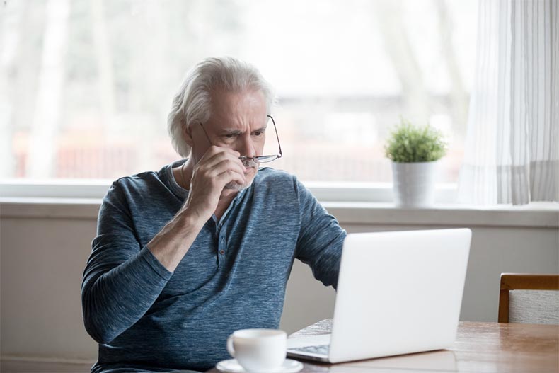 An older man sitting at a table and lowering his glasses as he skeptically looks at his laptop
