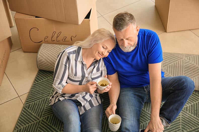 A mature couple with sitting on a carpet and drinking tea beside a stack of moving boxes