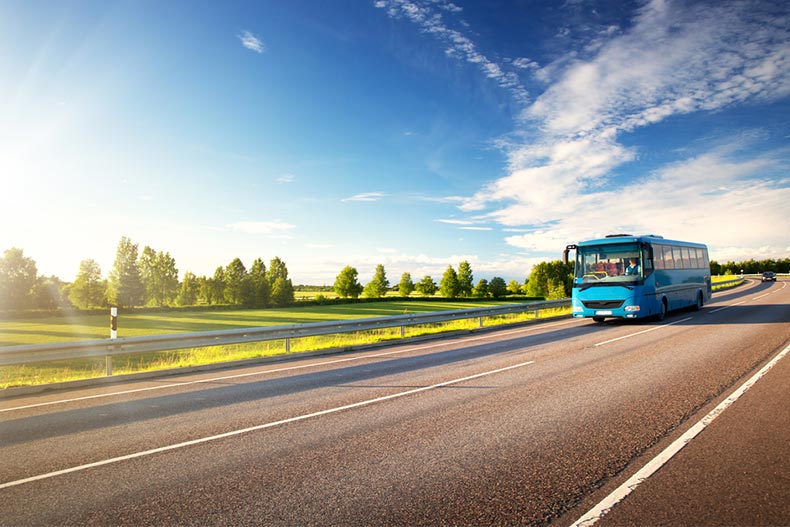 A blue bus driving down an asphalt road surrounded by green countryside