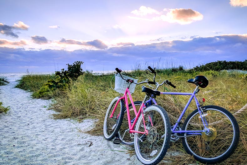 One pink and one purple bike parked between a sandy path and a grassy field, located in Fort Myers, Florida