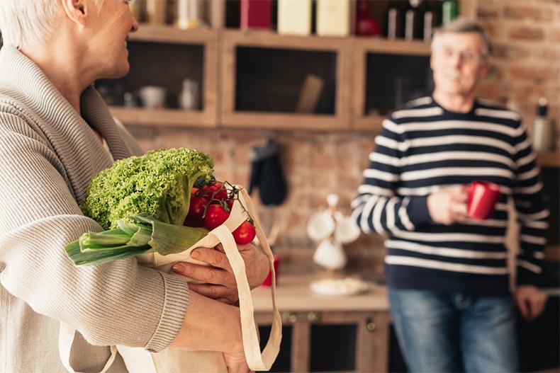 An elderly couple in their kitchen unpacking a reusable grocery bag filled with organic vegetables
