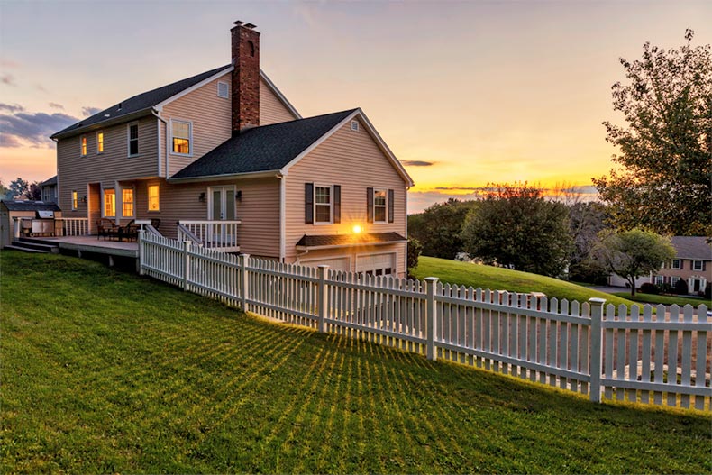 A white picket fence surrounding a beautiful colonial American house at sunset