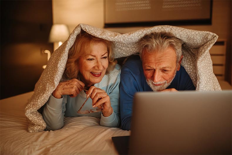An older couple on a bed under covers watching a movie