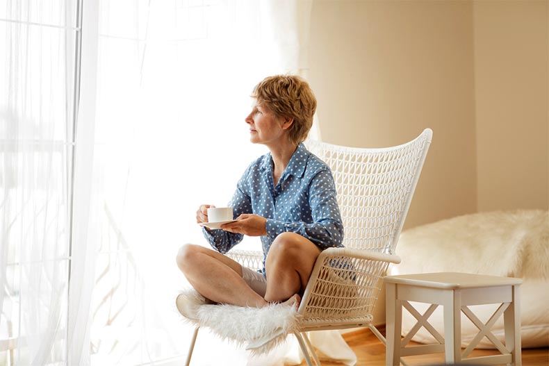 An active adult woman sitting on an armchair and looking out a window while drinking tea.