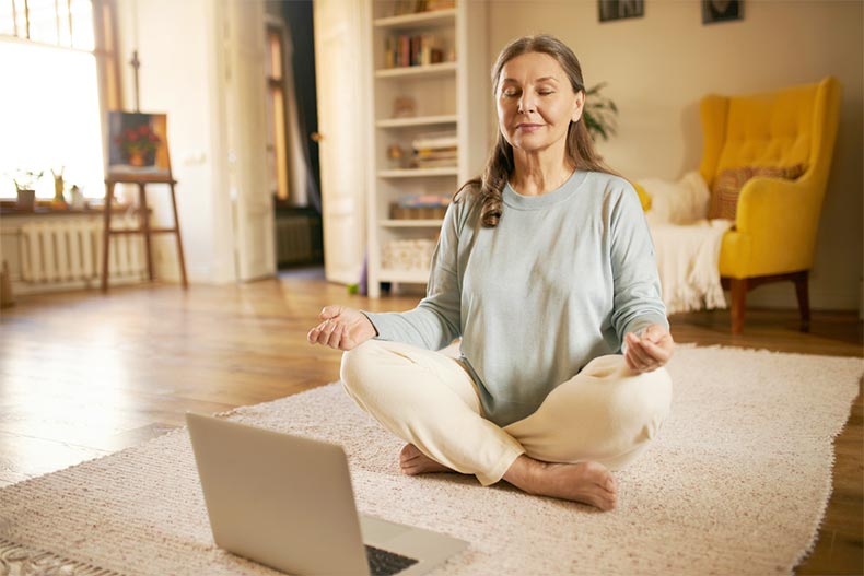 A senior woman sitting in front of an open laptop with her eyes closed and her legs crossed