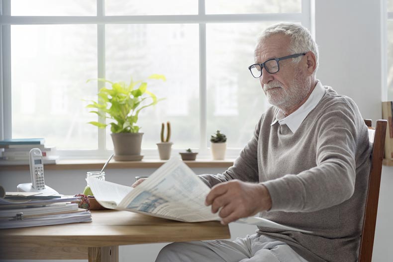 Senior man reading a newspaper at a kitchen table near a window