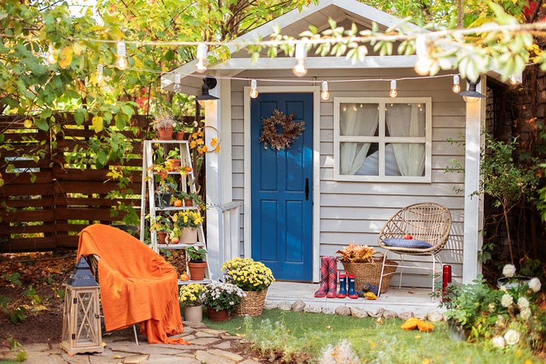 Exterior of a small, wooden guest house with a chair, a blanket, potted chrysanthemums, and pumpkins