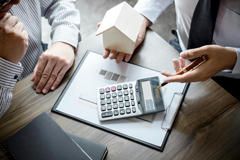 Closeup on the hands of a real estate agent holding a small wooden house and gesturing to a calculator