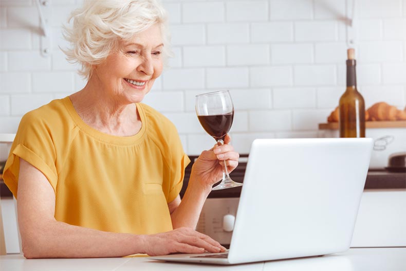 An older woman drinking wine while on a laptop in the kitchen