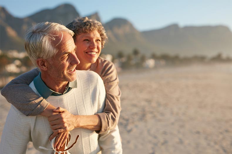 An older woman with her arms around her husband enjoying a sunset by the seashore