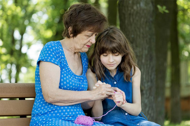 A grandmother teaching her granddaughter to crochet while sitting on a park bench in the summer