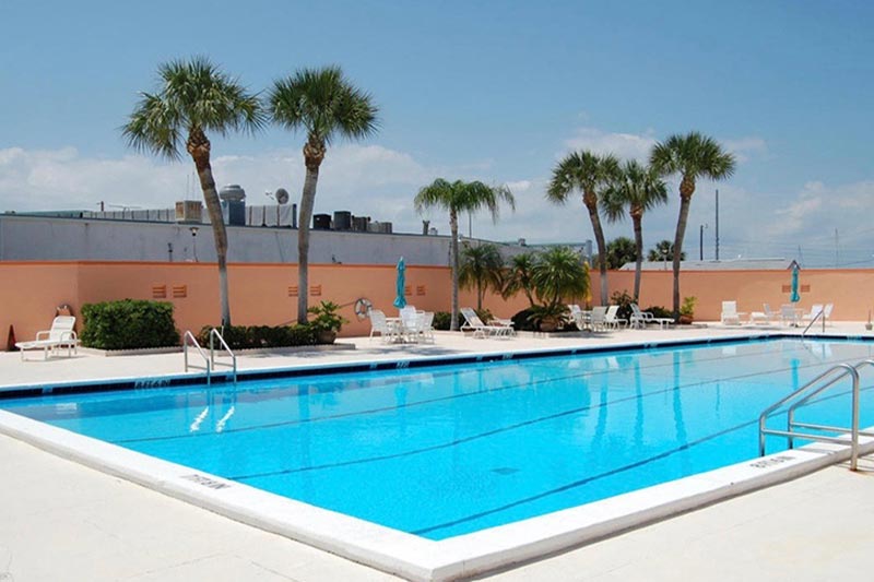 View of the outdoor pool and patio at Colonnades at Fort Pierce with palm trees and patio chairs on a sunny day.