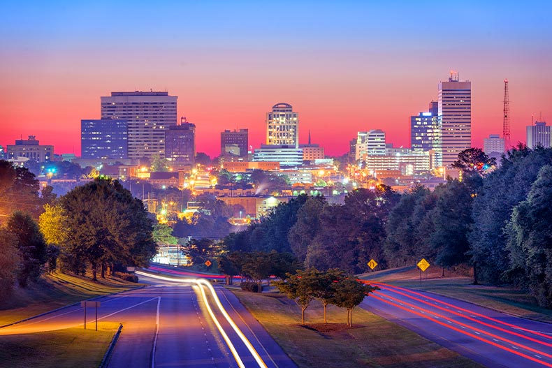 A wooded road with long-exposure light trails leading to the Columbia, South Carolina skyline at sunset