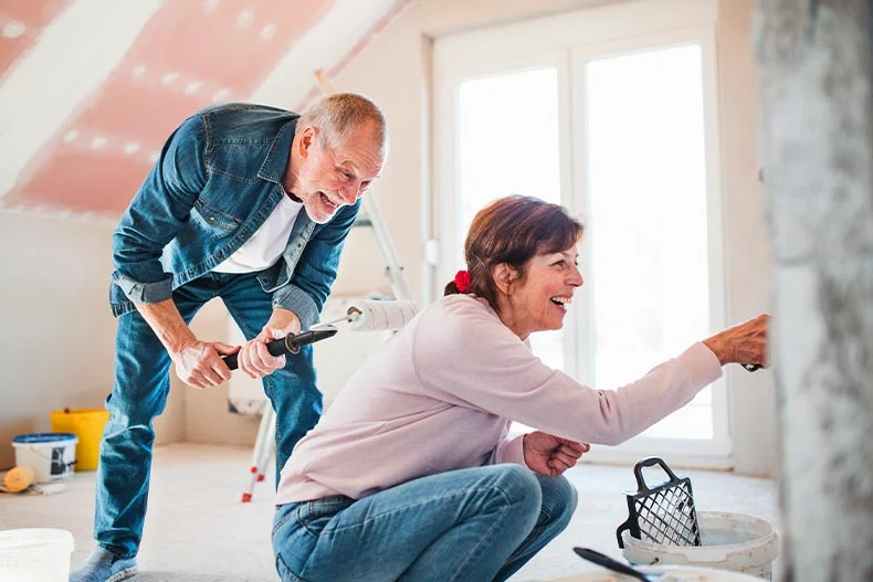 An elderly couple paints the walls of an attic while laughing