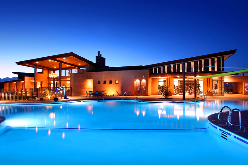 A blue, lighted, resort-style pool in front of the exterior of a clubhouse in Del Webb at Dove Mountain, located in Marana, Arizona at night