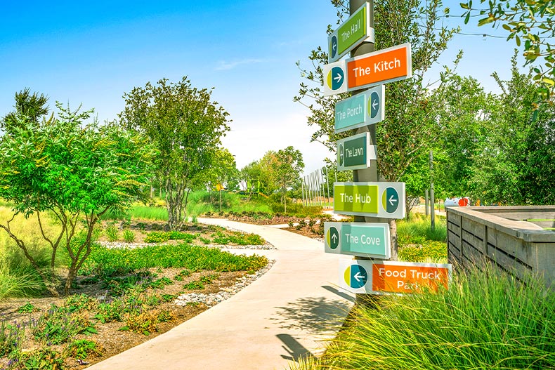 A pole with multicolored directional signs pointing to various amenities, located long a path with green plants in Del Webb at Union Park in Little Elm, Texas