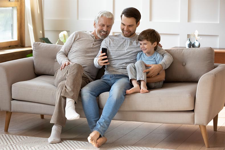 Young man sitting on a couch and smiling while using a mobile app with his elderly father and school-aged son