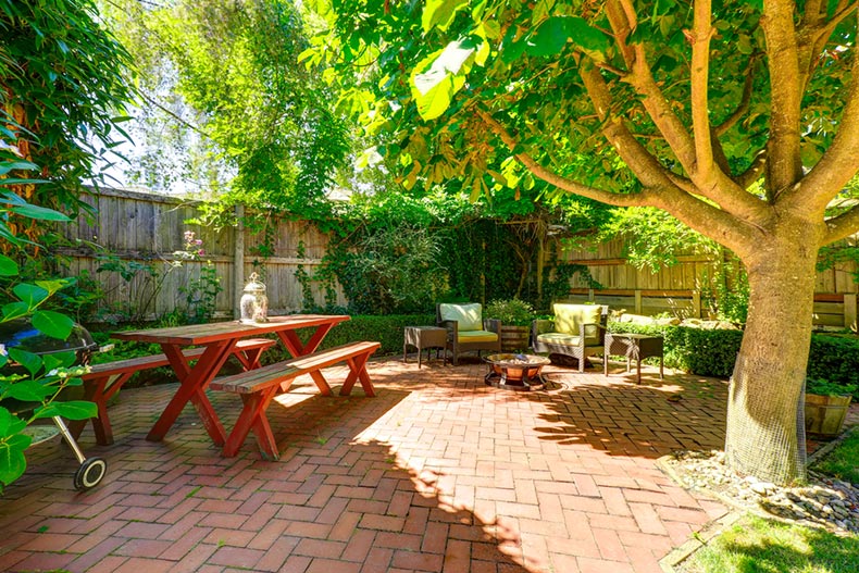 A backyard patio area with a wooden table and benches, wicker chairs, and a fire pit in a 55+ community