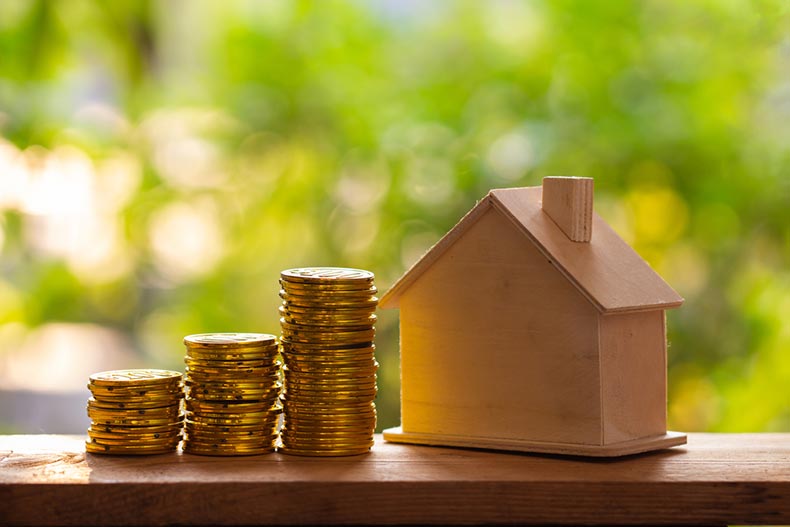 A small wooden model of a house beside three stacks of gold coins with a blurred nature background