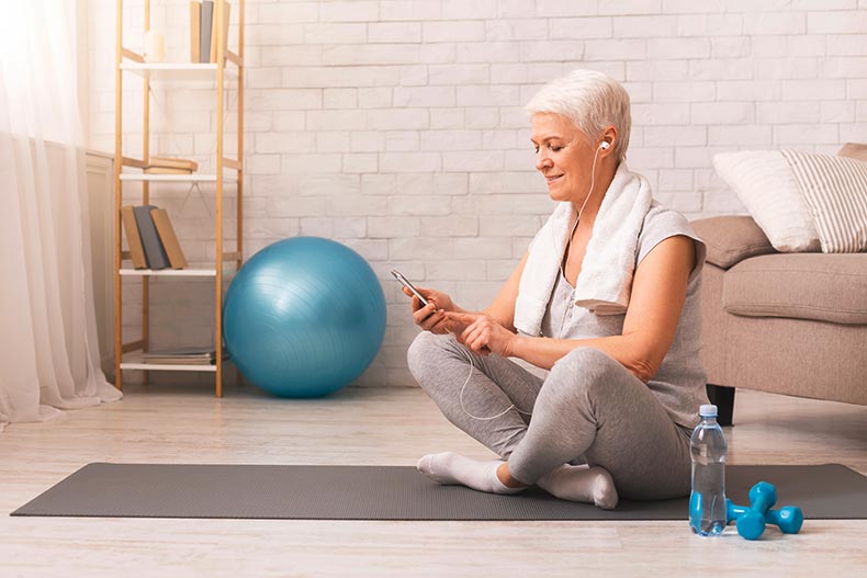 An older woman with a towel around her shoulders sitting on a yoga mat while selecting music on her smart phone