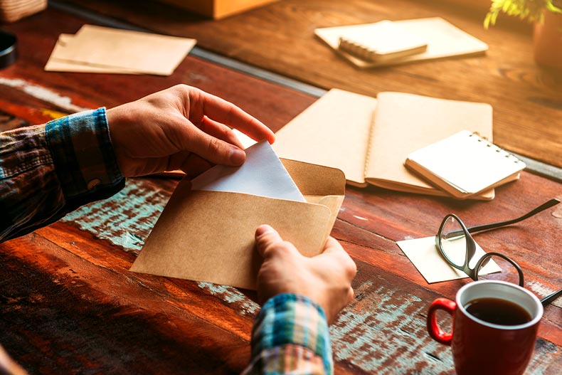 View of two hands opening a letter over a desk with a coffee cup, glasses, and several notebooks on top