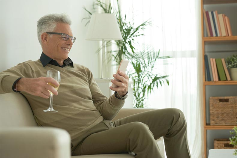 An older man smiling and video chatting on his phone while drinking wine