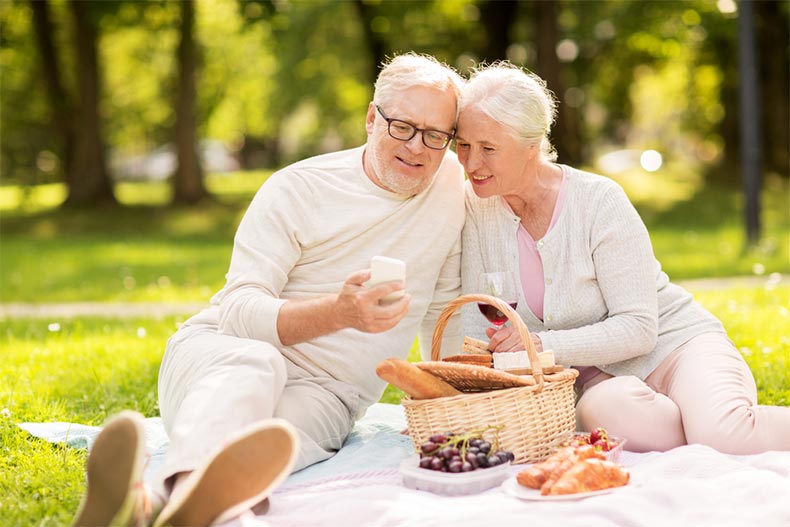 An older couple video chatting on a phone while having wine and a picnic in a park