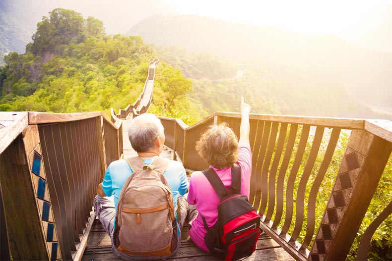Vacationing older couple sitting atop a long trail in green, mountainous landscape