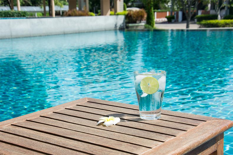 A glass of cold water with ice cubes and a slice of lime on wooden table by a swimming pool