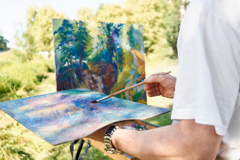 Closeup of a senior man's hands holding a painting brush and a palette while painting in a park