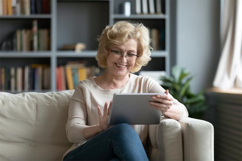 An older woman smiling while sitting on a couch and browsing homes for sale on a tablet