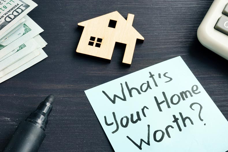 A wooden house cutout on a table beside a pen and a note reading "What's Your Home Worth?"