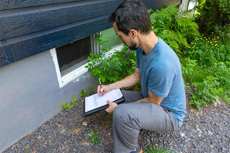 A home inspector writing notes while he inspects the outside of a basement window