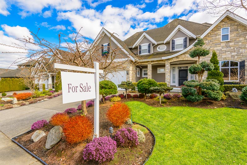 A "For Sale" sign in front of a luxurious house with a landscaped yard