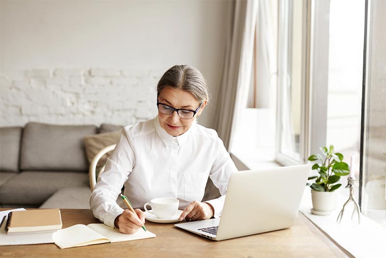 An older woman making notes in a notebook while working on a laptop and drinking coffee