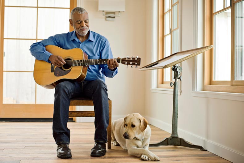 A 55+ man playing guitar while his Labrador Retriever lays at his feet