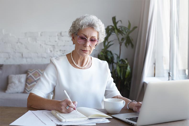 An older woman with glasses writing notes as she uses her laptop to take an online course
