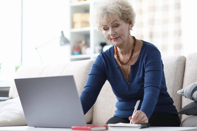 An older woman writing notes as she uses her laptop to take an online course