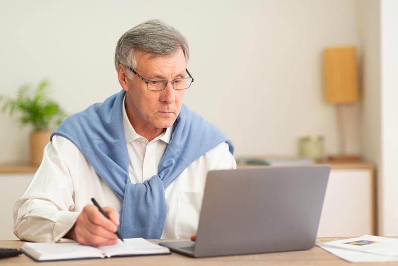 An older man writing notes while taking an online course on his laptop