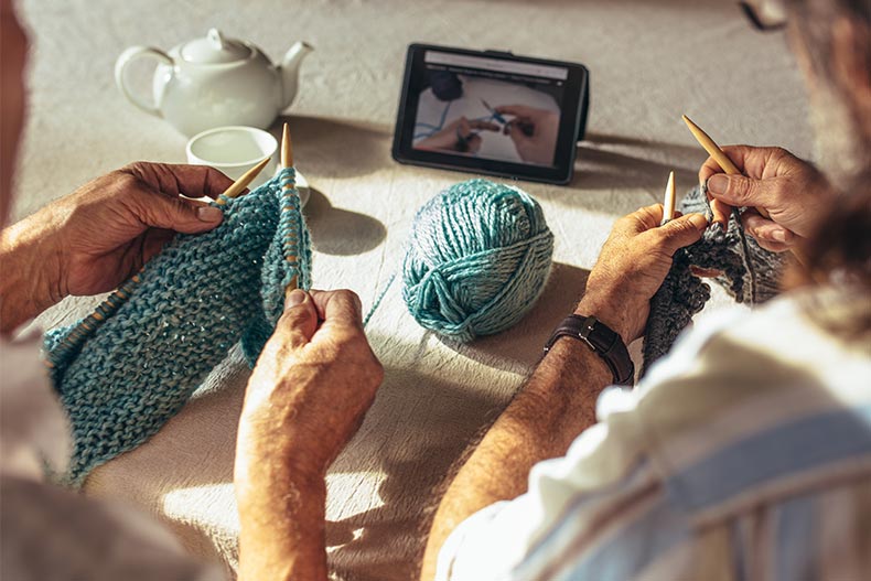 Hands of senior men holding knitting needles and wool yard while sitting at table and learning knitting from online videos