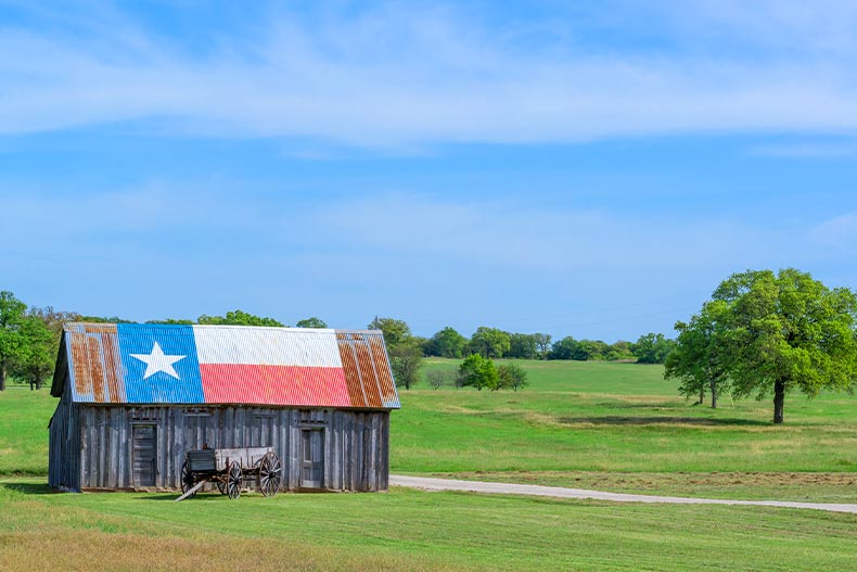 Exterior view of an old barn and buggy with a Texas state flag painted on the roof, located in the middle of a prairie
