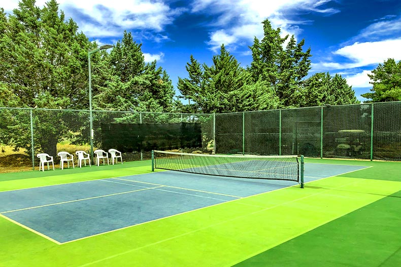 A blue and green pickleball court with five white lawn chairs on the sidelines and pine trees outside the gate, located in Villages of Lynx Creek in Dewey, Arizona