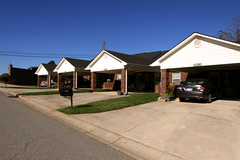 A row of four brown brick attached homes with white and black roofs with cars parked in the driveways, located in Magnolia Place in Lowell, North Carolina