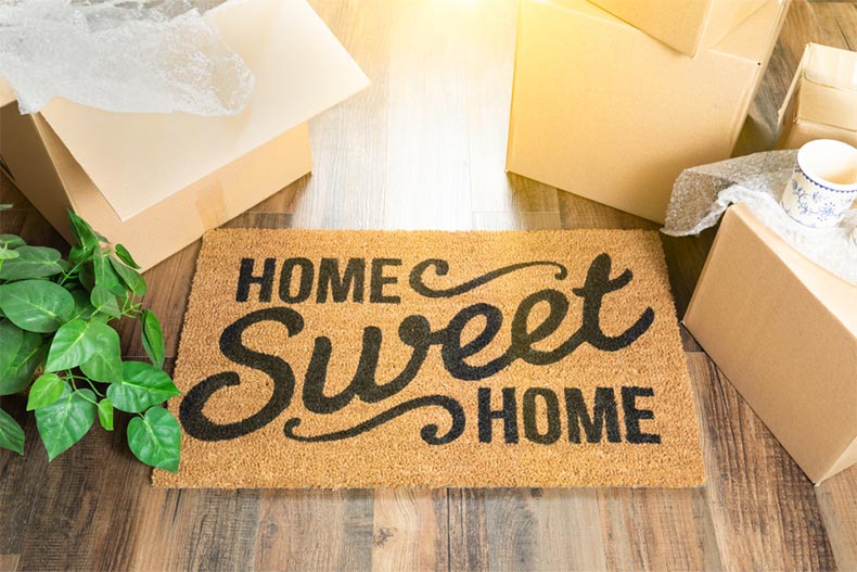 A "Home Sweet Home" welcome mat surrounded by moving boxes on a hardwood floor
