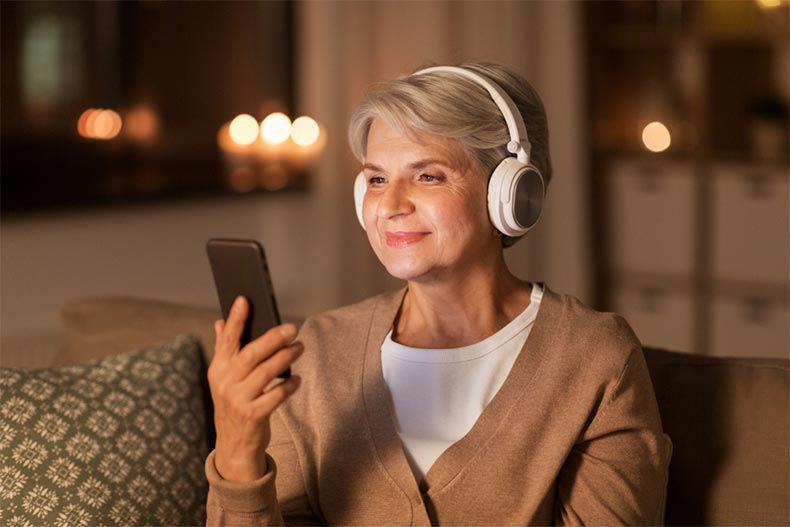 A senior woman wearing headphones and smiling while listening to music on a smartphone at home in the evening