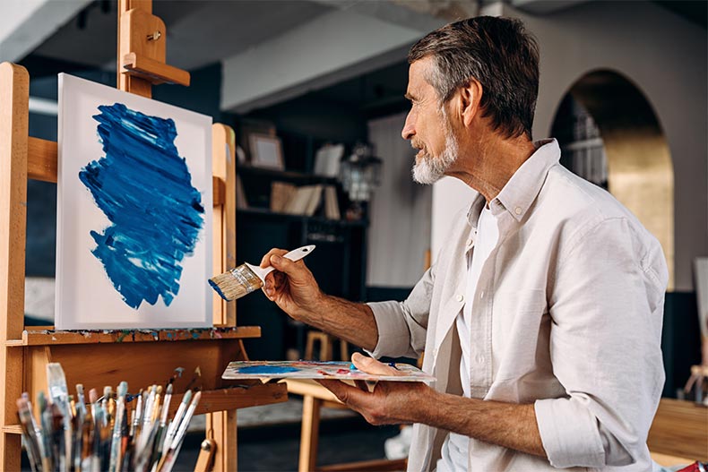 A male artist sitting in front of easel and painting an abstract picture with a large paintbrush