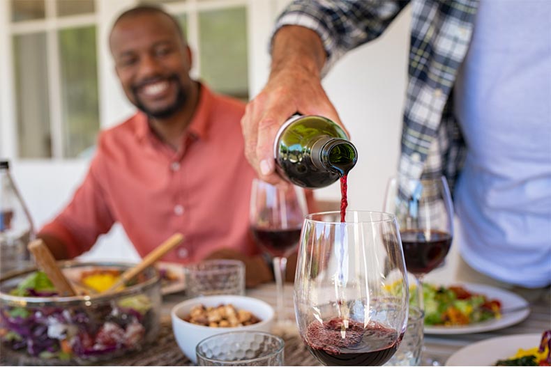 A man watching his older friend poor a glass of red wine