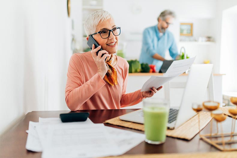 An older woman calculating finances while an older man cooks in the kitchen