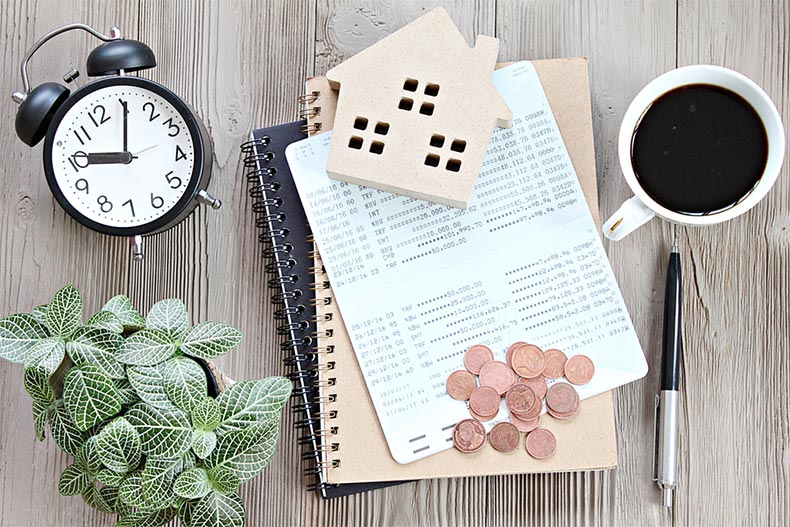 A clock, a cup of coffee, and a wooden house silhouette on top of home mortgage calculations